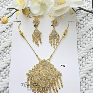 May include: Gold necklace and earrings set with clear crystal stones. The necklace features a large pendant with a floral design and dangling strands of crystals. The earrings are also floral shaped with dangling crystals. The set is labeled "N11".