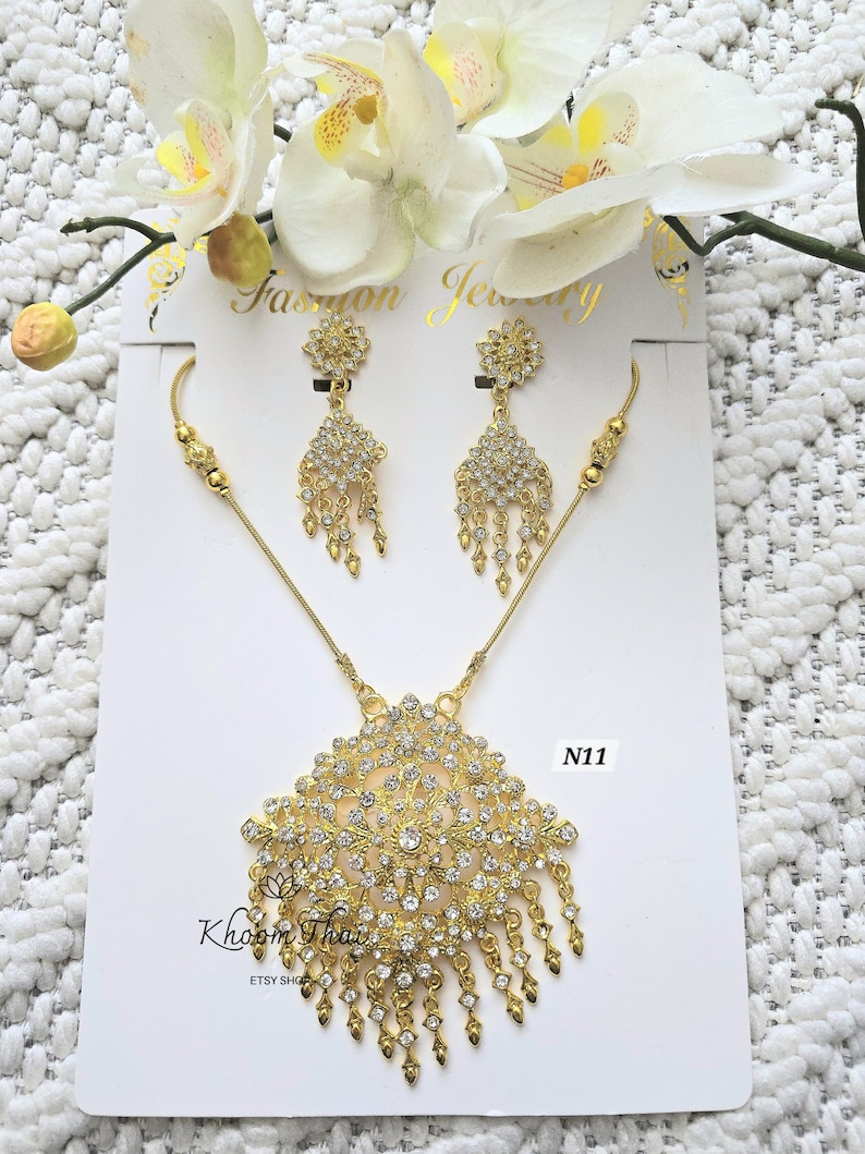May include: Gold necklace and earrings set with clear crystal stones. The necklace features a large pendant with a floral design and dangling strands of crystals. The earrings are also floral shaped with dangling crystals. The set is labeled "N11".