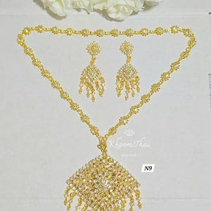 May include: A gold necklace with a square pendant and matching earrings. The pendant and earrings are adorned with clear rhinestones. The necklace is labeled "Khoon Thai Etsy Shop N9".