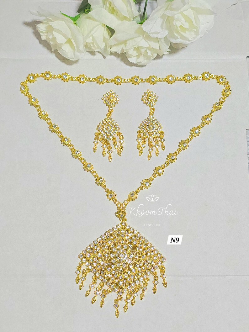 May include: A gold necklace with a square pendant and matching earrings. The pendant and earrings are adorned with clear rhinestones. The necklace is labeled "Khoon Thai Etsy Shop N9".