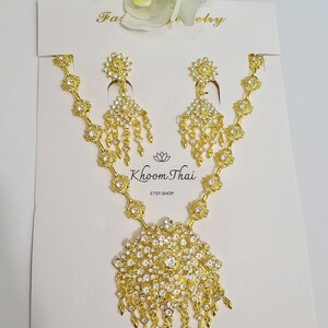 May include: A gold necklace and earrings set with a large, round pendant adorned with clear rhinestones. The pendant has a dangling fringe of smaller rhinestones. The set is displayed on a white card with the text "Khoon Thai ETSY SHOP" and "N7".