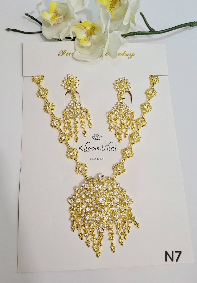 May include: A gold necklace and earrings set with a large, round pendant adorned with clear rhinestones. The pendant has a dangling fringe of smaller rhinestones. The set is displayed on a white card with the text "Khoon Thai ETSY SHOP" and "N7".