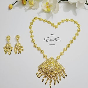 May include: A gold necklace and earrings set with a large pendant and dangling drops. The pendant is adorned with clear rhinestones and has a pointed design. The necklace is made of a chain with small flower-shaped links. The earrings are also adorned with clear rhinestones. The set is labeled "Khoon Thai Etsy Shop" and "N6".