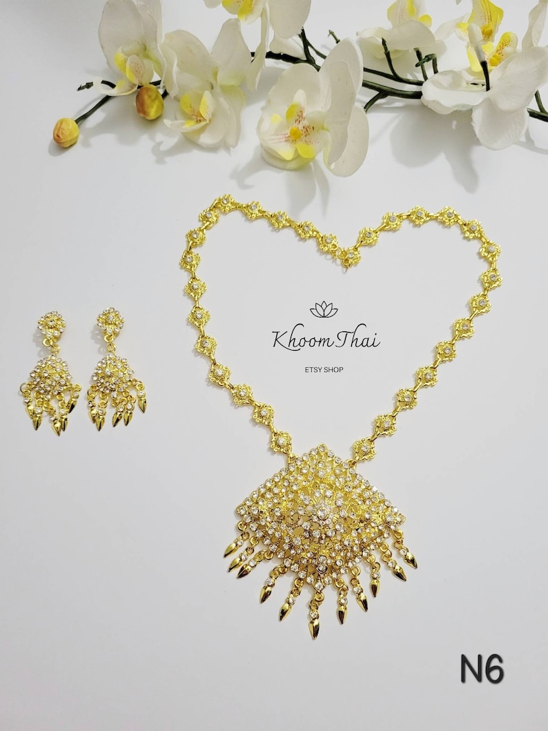 May include: A gold necklace and earrings set with a large pendant and dangling drops. The pendant is adorned with clear rhinestones and has a pointed design. The necklace is made of a chain with small flower-shaped links. The earrings are also adorned with clear rhinestones. The set is labeled "Khoon Thai Etsy Shop" and "N6".
