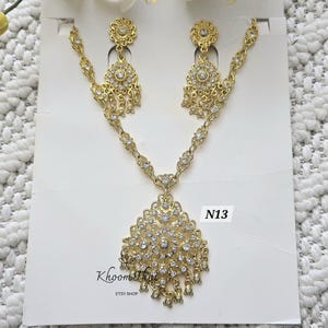 May include: Gold necklace and earrings set with clear rhinestone accents. The necklace features a large pendant with a floral design. The earrings are dangle style with a similar floral design. The set is labeled "N13" and "KhoomaThai Etsy Shop".