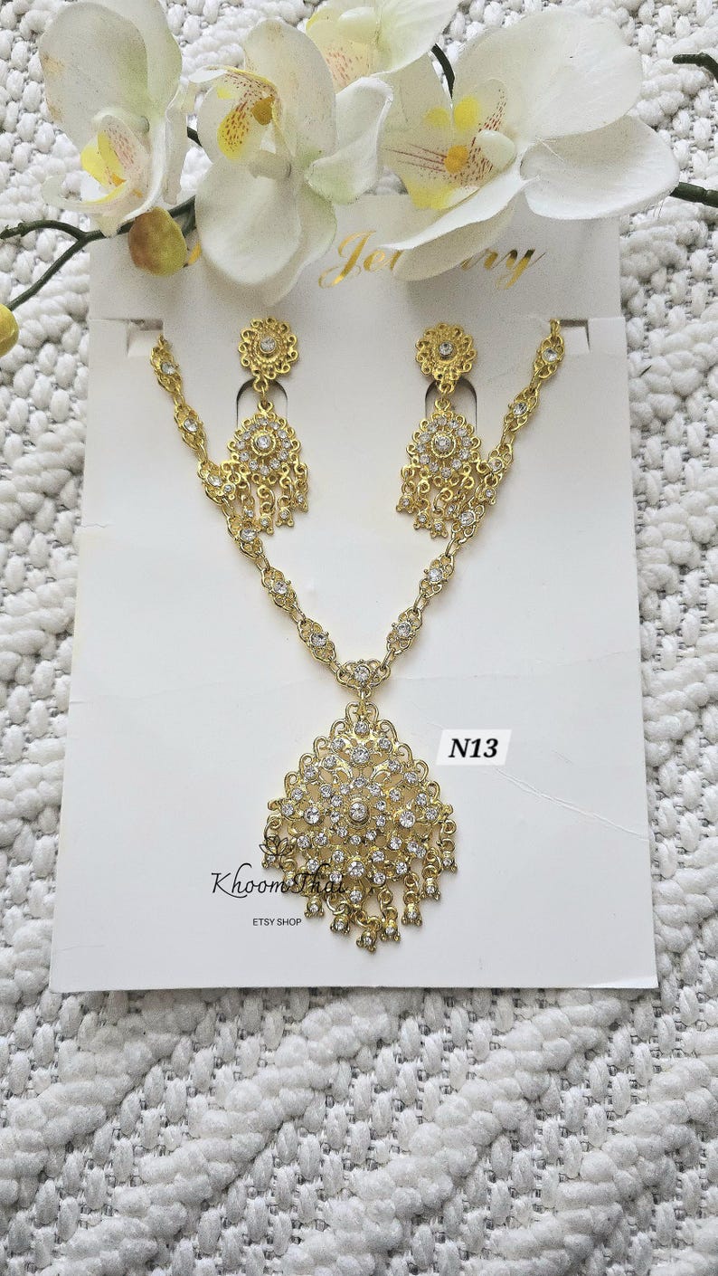 May include: Gold necklace and earrings set with clear rhinestone accents. The necklace features a large pendant with a floral design. The earrings are dangle style with a similar floral design. The set is labeled "N13" and "KhoomaThai Etsy Shop".