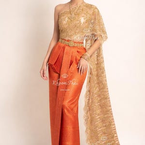May include: A traditional Thai dress in shades of gold and orange. The one-shoulder top and shawl are adorned with intricate lace. The skirt is a vibrant orange with a textured pattern, and the waist is cinched with a gold belt. The dress is from Khoom Thai Etsy Shop.