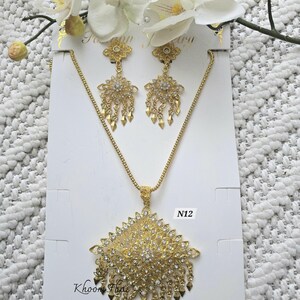 May include: A gold-tone necklace and earrings set with a large, ornate pendant and dangling earrings. The pendant and earrings are covered in sparkling clear rhinestones. The set is labeled "N12" and "Khoon Thai Etsy Shop".