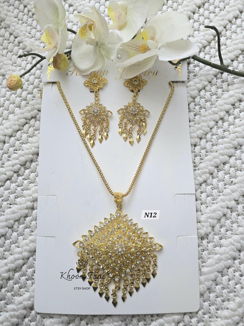 May include: A gold-tone necklace and earrings set with a large, ornate pendant and dangling earrings. The pendant and earrings are covered in sparkling clear rhinestones. The set is labeled "N12" and "Khoon Thai Etsy Shop".