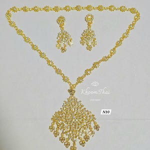 May include: A gold necklace with a large, diamond-shaped pendant and matching earrings. The pendant and earrings are adorned with clear rhinestones. The necklace is labeled "N10" and "Khoon Thai Etsy Shop".