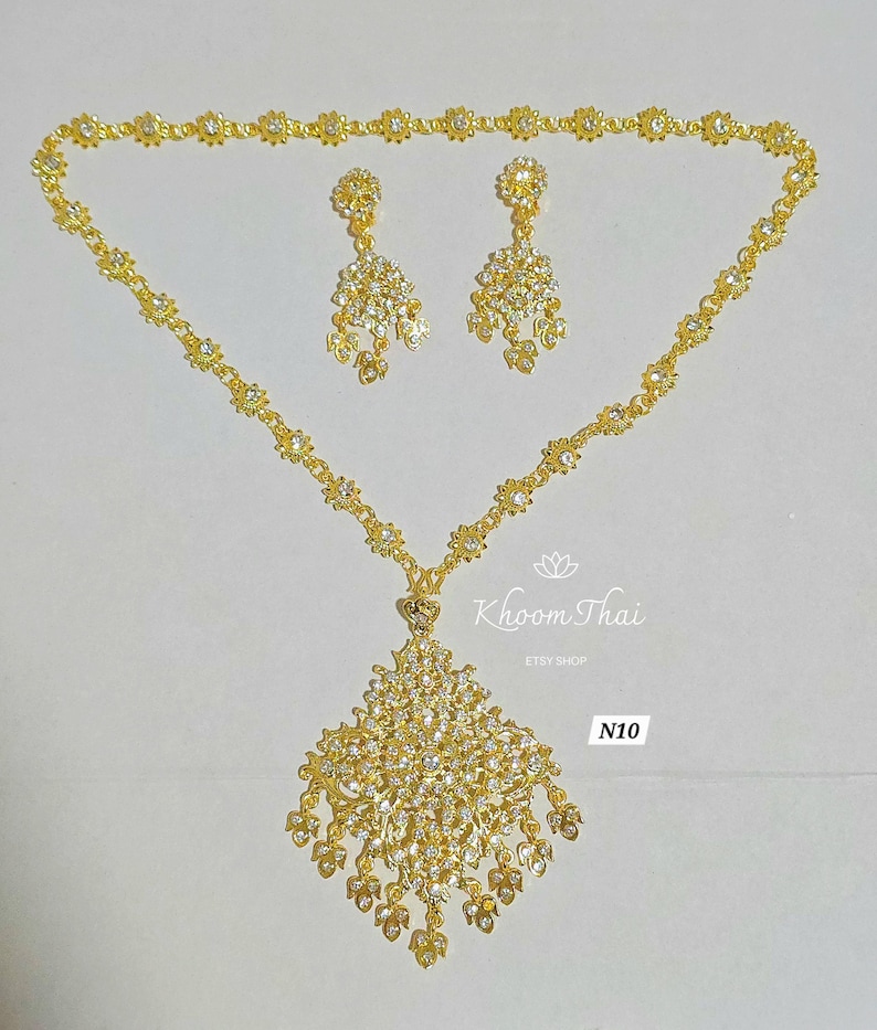 May include: A gold necklace with a large, diamond-shaped pendant and matching earrings. The pendant and earrings are adorned with clear rhinestones. The necklace is labeled "N10" and "Khoon Thai Etsy Shop".