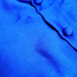 May include: Close-up of a bright blue fabric with two buttons. The fabric has a subtle texture and a smooth, silky feel.