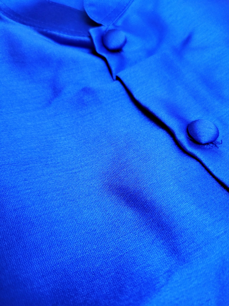 May include: Close-up of a bright blue fabric with two buttons. The fabric has a subtle texture and a smooth, silky feel.