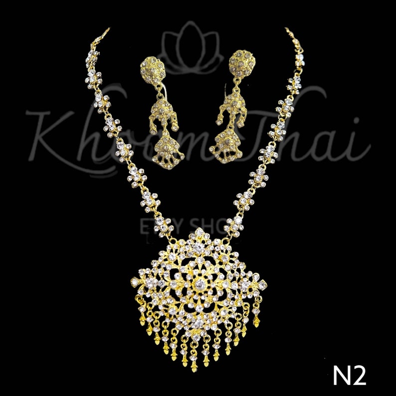May include: A gold necklace and earrings set with a large, ornate pendant and dangling drops of clear rhinestones. The pendant is a circular design with a floral pattern. The necklace and earrings are made of metal and have a shiny finish.