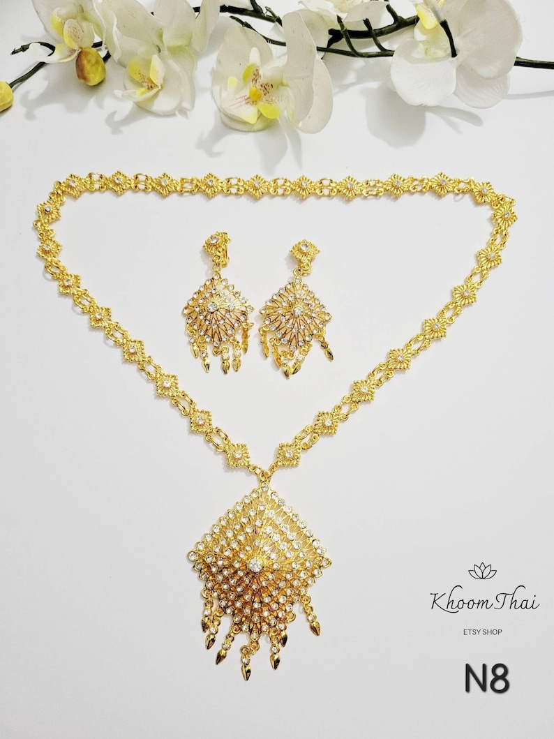 May include: A gold necklace with a large, ornate pendant and matching earrings. The pendant and earrings are decorated with clear rhinestones. The necklace is on a white background. N8