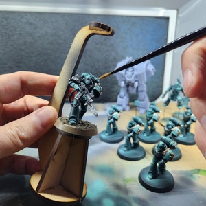 May include: A teal-coloured miniature space marine figure holding a gun, standing on a wooden base. The figure is being painted with a brush by a person's hand. There are other miniature figures in the background.