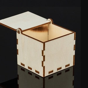 Op de afbeelding: Een kleine, open houten doos met een scharnierend deksel. De doos is gemaakt van lichtgekleurd hout met donkerbruine randen en verbindingen. Het deksel staat iets open en onthult de lege binnenkant. De doos is waarschijnlijk voor het opbergen van kleine items.