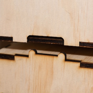 Puede incluir: Dos cajas de madera con bordes negros apiladas una encima de la otra. La caja superior tiene un peque&ntilde;o recorte en el borde delantero. La caja inferior tiene dos peque&ntilde;os recortes en el borde delantero.