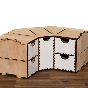 Puede incluir: Una estanter&iacute;a de esquina de madera con seis cajones. Los cajones son blancos con un borde negro. La estanter&iacute;a est&aacute; hecha de madera clara con un acabado natural.