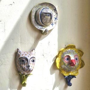May include: Three ceramic wall hooks featuring hand-painted designs. The top hook depicts a crescent moon face with blue stars. The bottom left hook shows a cat face with brown spots. The bottom right hook displays a yellow flower face with a red beak.