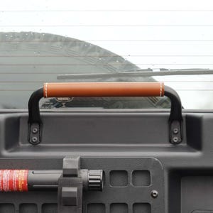 May include: A black metal grab handle with a brown leather grip and white stitching. The handle is mounted on a black surface with visible mounting hardware. The handle is designed for a vehicle.