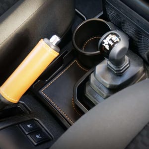 May include: Interior car detail featuring a tan leather-wrapped handbrake and a black leather-trimmed console. The console includes a cup holder and a gear shift knob. The stitching on the leather is a contrasting yellow.