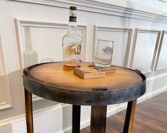 May include: A rustic, round side table made from a repurposed wooden barrel. The table features a dark metal band around the top and a lower shelf. A bottle of whiskey, a glass, and coasters are displayed on the table.