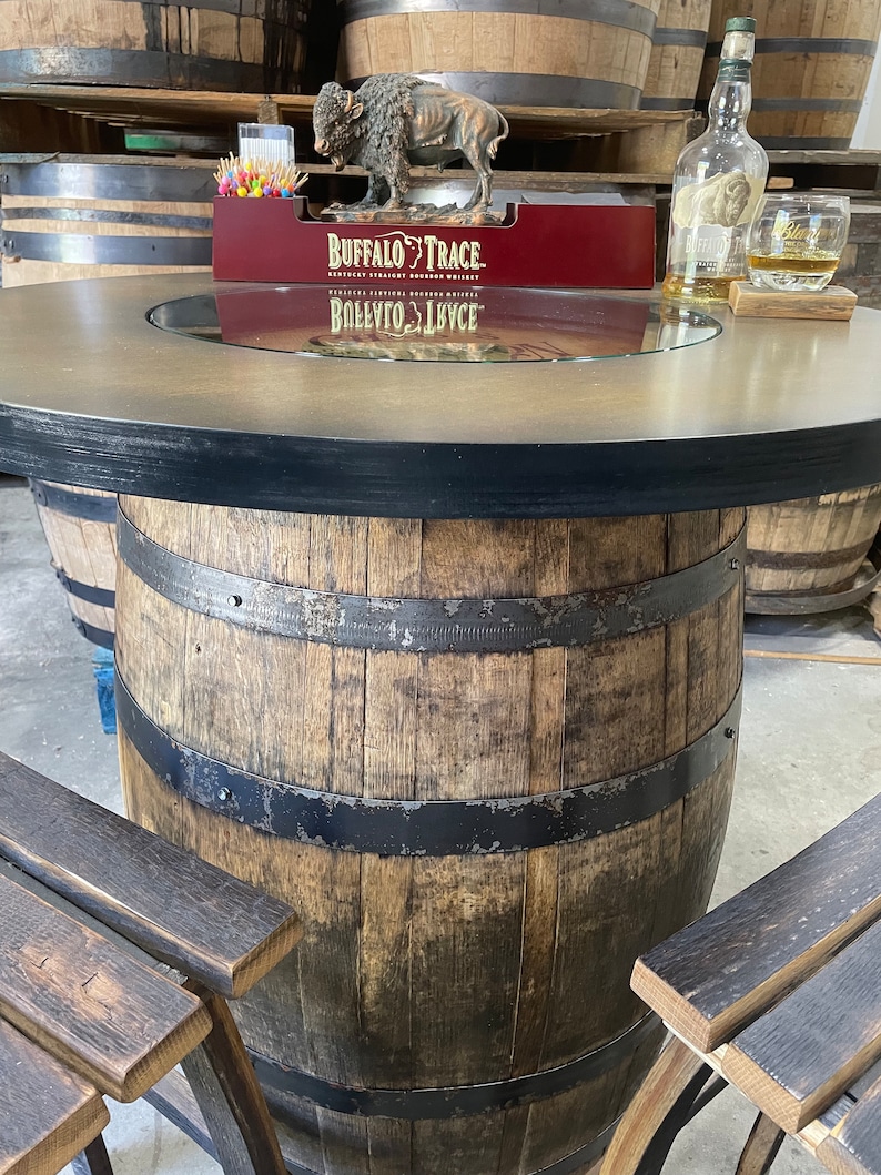 Puede incluir: Una mesa de barril de madera con una tapa de vidrio y una banda de metal negro alrededor de la parte superior. La mesa est&aacute; rodeada de barriles de madera y una peque&ntilde;a estatua de b&uacute;falo de metal est&aacute; encima de la mesa. La mesa tiene un letrero que dice "Buffalo Trace Kentucky Straight Bourbon Whiskey" y "Ballito Lycee".