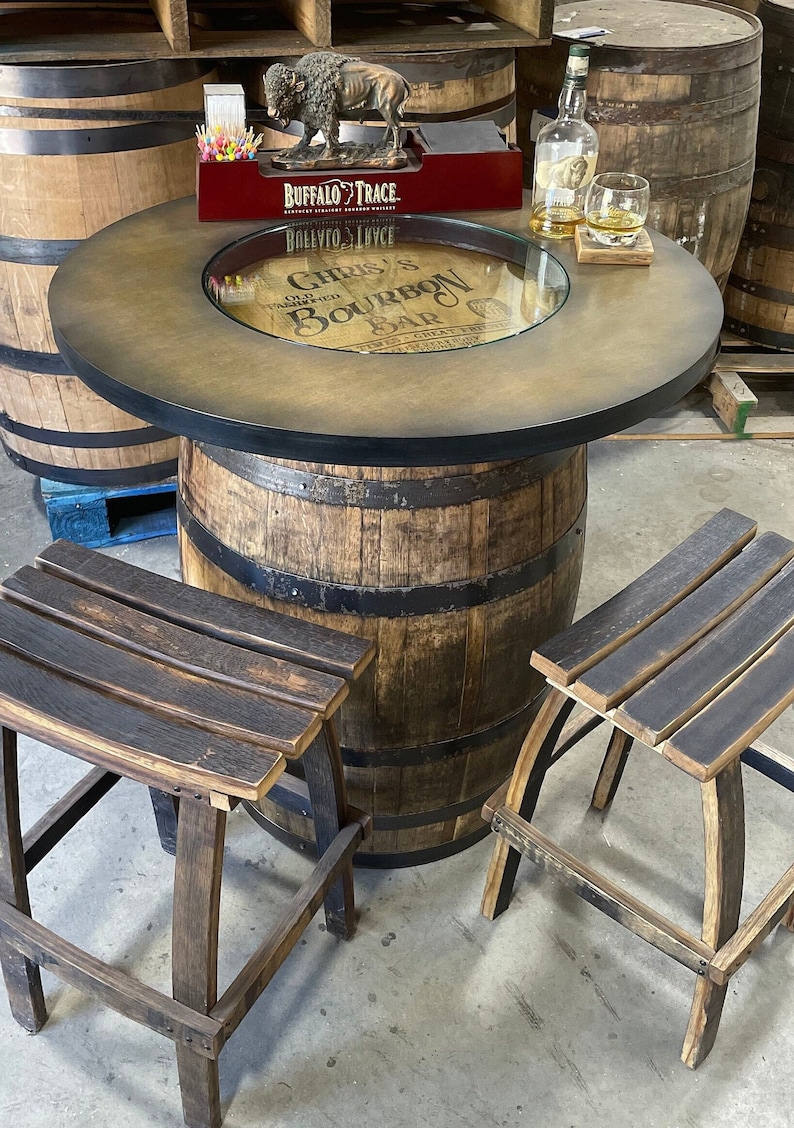 Puede incluir: Una mesa de barril de madera con una tapa de vidrio y un acabado marr&oacute;n desgastado. La mesa tiene una banda de metal negro alrededor de la parte superior e inferior. Dos taburetes de barril de madera con asientos te&ntilde;idos de negro se colocan junto a la mesa. La tapa de la mesa tiene una etiqueta que dice "Chris's Old Fashioned Bourbon Bar".