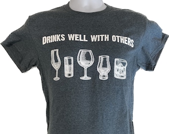 May include: Dark gray t-shirt with the phrase "DRINKS WELL WITH OTHERS" in white block letters. Below the text are white line drawings of five different types of glasses. The shirt is displayed on a white mannequin.