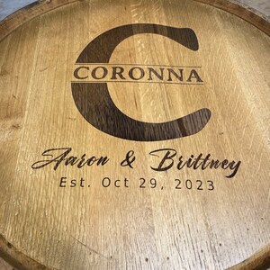Puede incluir: Una tapa de barril de madera con una inscripci&oacute;n personalizada. La inscripci&oacute;n dice "Coronna Aaron & Brittney Est. Oct 29, 2023".