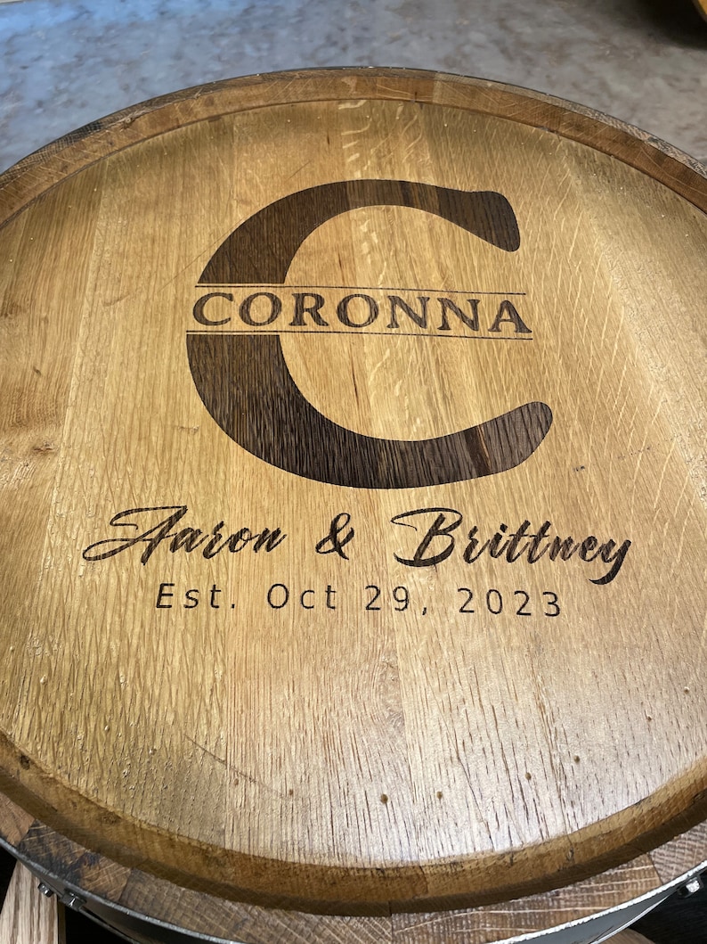Puede incluir: Una tapa de barril de madera con una inscripci&oacute;n personalizada. La inscripci&oacute;n dice "Coronna Aaron & Brittney Est. Oct 29, 2023".
