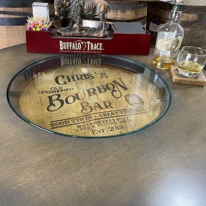 Puede incluir: Una tapa de barril de madera con un inserto de vidrio, que presenta el texto "Chris's Bourbon Bar" y las palabras "Good Times - Great Friends" y "Where everybody gets a second shot". La tapa del barril est&aacute; sobre una mesa marr&oacute;n oscura.