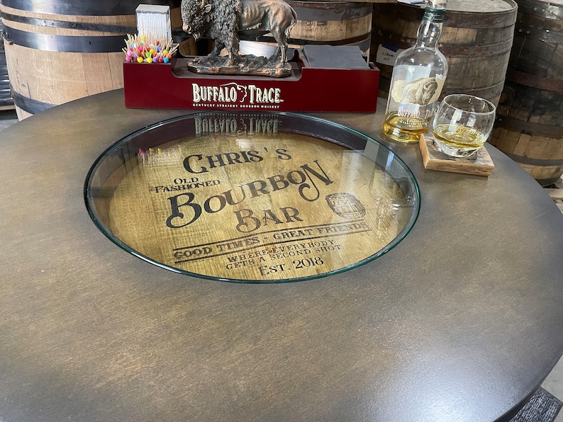 Puede incluir: Una tapa de barril de madera con un inserto de vidrio, que presenta el texto "Chris's Bourbon Bar" y las palabras "Good Times - Great Friends" y "Where everybody gets a second shot". La tapa del barril est&aacute; sobre una mesa marr&oacute;n oscura.
