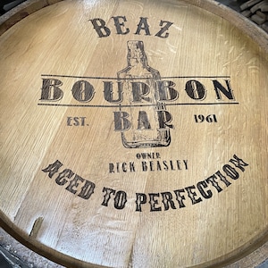 Puede incluir: Un barril de madera con el texto "BEAZ BOURBON BAR EST. 1961 OWNER RICK BEASLEY AGED TO PERFECTION" quemado en la madera.