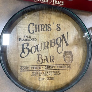 Puede incluir: Un letrero redondo de madera con marco negro, que presenta el texto "Chris's Old Fashioned Bourbon Bar", "Good Times - Great Friends", "Where Everybody Gets A Second Shot" y "Est. 2018".