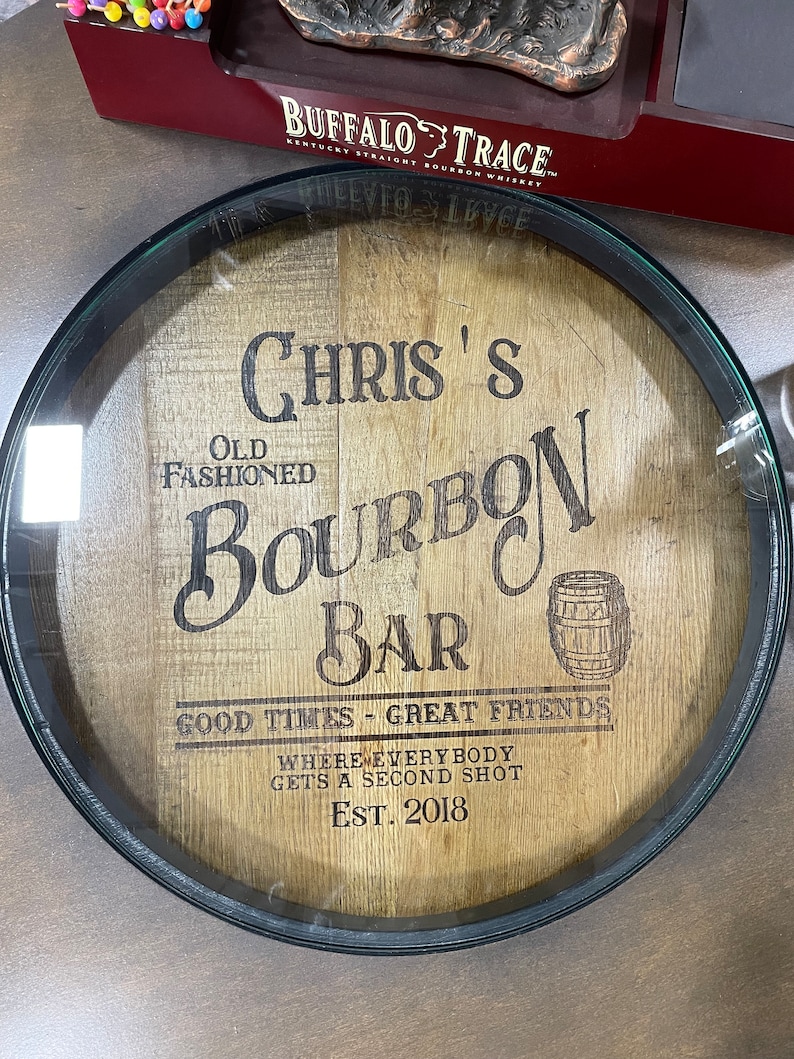 Puede incluir: Un letrero redondo de madera con marco negro, que presenta el texto "Chris's Old Fashioned Bourbon Bar", "Good Times - Great Friends", "Where Everybody Gets A Second Shot" y "Est. 2018".
