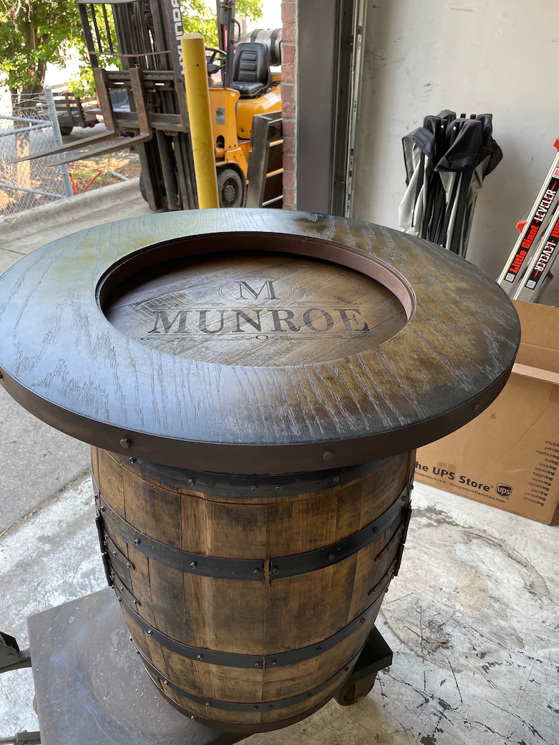 Puede incluir: Una mesa de barril de madera con una tapa de madera marr&oacute;n oscuro y un borde de metal negro. La tapa tiene un recorte circular con el nombre "M. MUNROE" grabado en la madera.