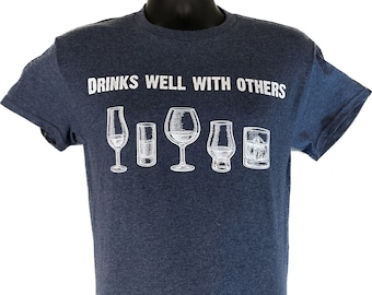 May include: A navy blue t-shirt with the text "DRINKS WELL WITH OTHERS" above a row of five different drink glasses in white. The glasses include a champagne flute, a shot glass, a wine glass, a snifter, and an old-fashioned glass.