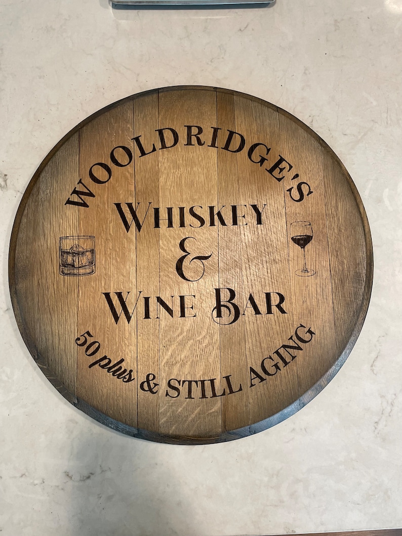 Puede incluir: Un letrero redondo de madera con el texto "Wooldridge's Whiskey & Wine Bar 50 plus & Still Aging" en letras negras. El letrero presenta una peque&ntilde;a ilustraci&oacute;n de un vaso de whisky y un vaso de vino.
