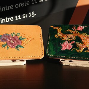 May include: Two leather wallets with hand-painted designs. One wallet is tan with a floral design in shades of pink, purple, and green. The other wallet is green with a gold fish and pink flowers.