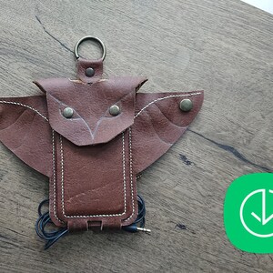 Könnte beinhalten: Eine braune Lederhülle für ein Handy in Form einer Eule mit einem Loch für Kopfhörer. Die Hülle hat eine Klappe, die mit einem Druckknopf geschlossen wird.