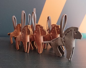 May include: A group of brown and gray leather horse ornaments with a hanging loop. The ornaments are made of stitched leather and have a rustic look.