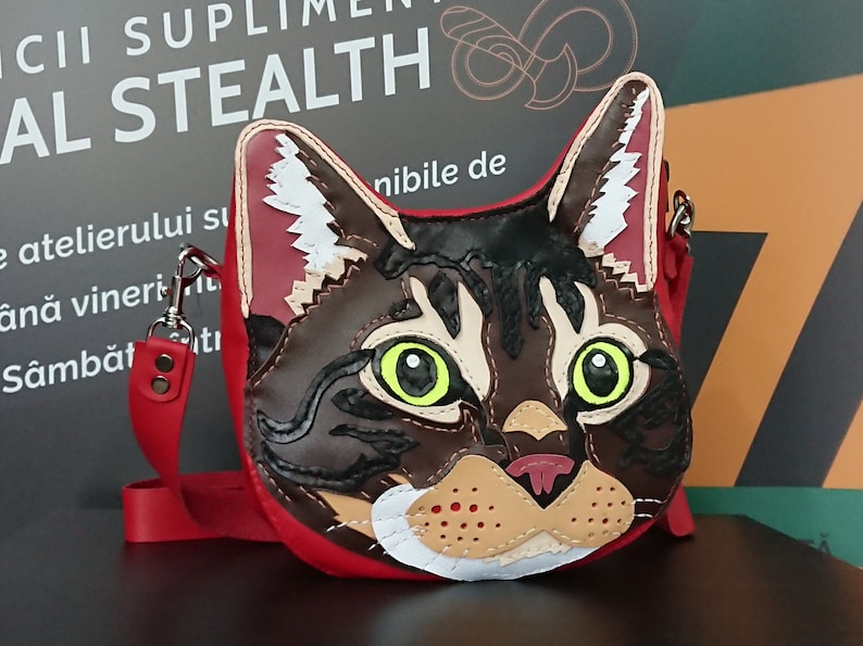 May include: A red leather crossbody bag with a cat face design. The cat has brown fur, green eyes, and a pink nose. The bag has a red strap and a silver clasp.