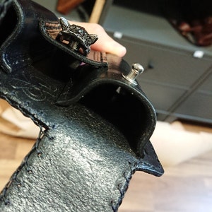 以下が含まれることがあります： 黒い革の財布。装飾的なオオカミの頭の留め金が付いています。財布は質感のある表面で、端にステッチが見えます。シルバーのスナップ留めも確認できます。