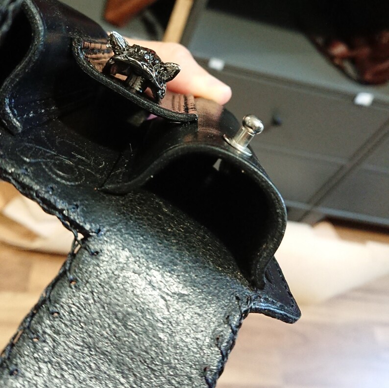 以下が含まれることがあります： 黒い革の財布。装飾的なオオカミの頭の留め金が付いています。財布は質感のある表面で、端にステッチが見えます。シルバーのスナップ留めも確認できます。