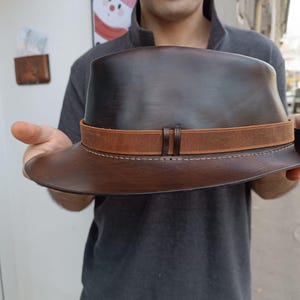 Może przedstawiać: Brązowy sk&oacute;rzany kapelusz fedora z brązowym sk&oacute;rzanym paskiem i szwami. Kapelusz ma szeroki rondel.