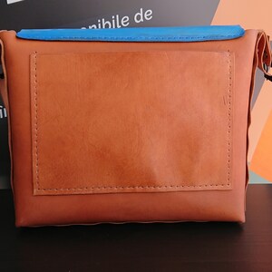 May include: A brown leather crossbody bag with a blue top flap and an orange tag. The bag has a rectangular design with a stitched pocket on the front. The bag has a strap and a buckle.