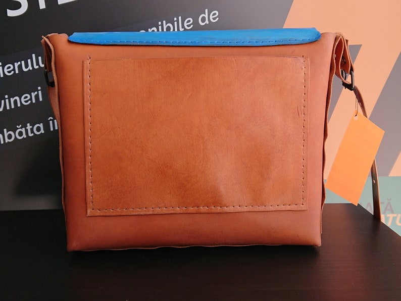 May include: A brown leather crossbody bag with a blue top flap and an orange tag. The bag has a rectangular design with a stitched pocket on the front. The bag has a strap and a buckle.