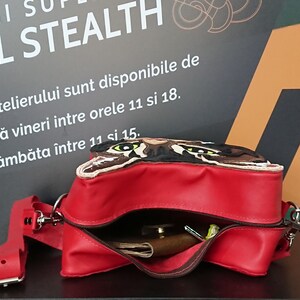 May include: A red leather crossbody bag with a flap featuring an embroidered cat face. The bag is open, revealing a brown leather wallet and other items inside.